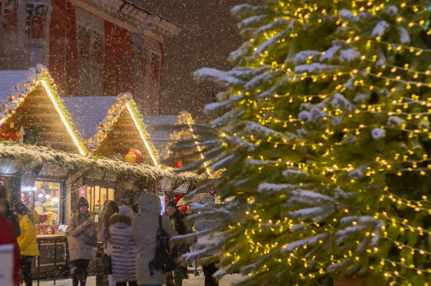 Weihnachtsmarkt-Hütten mit Lichterketten neben einem beleuchteten Tannenbaum im Schnee