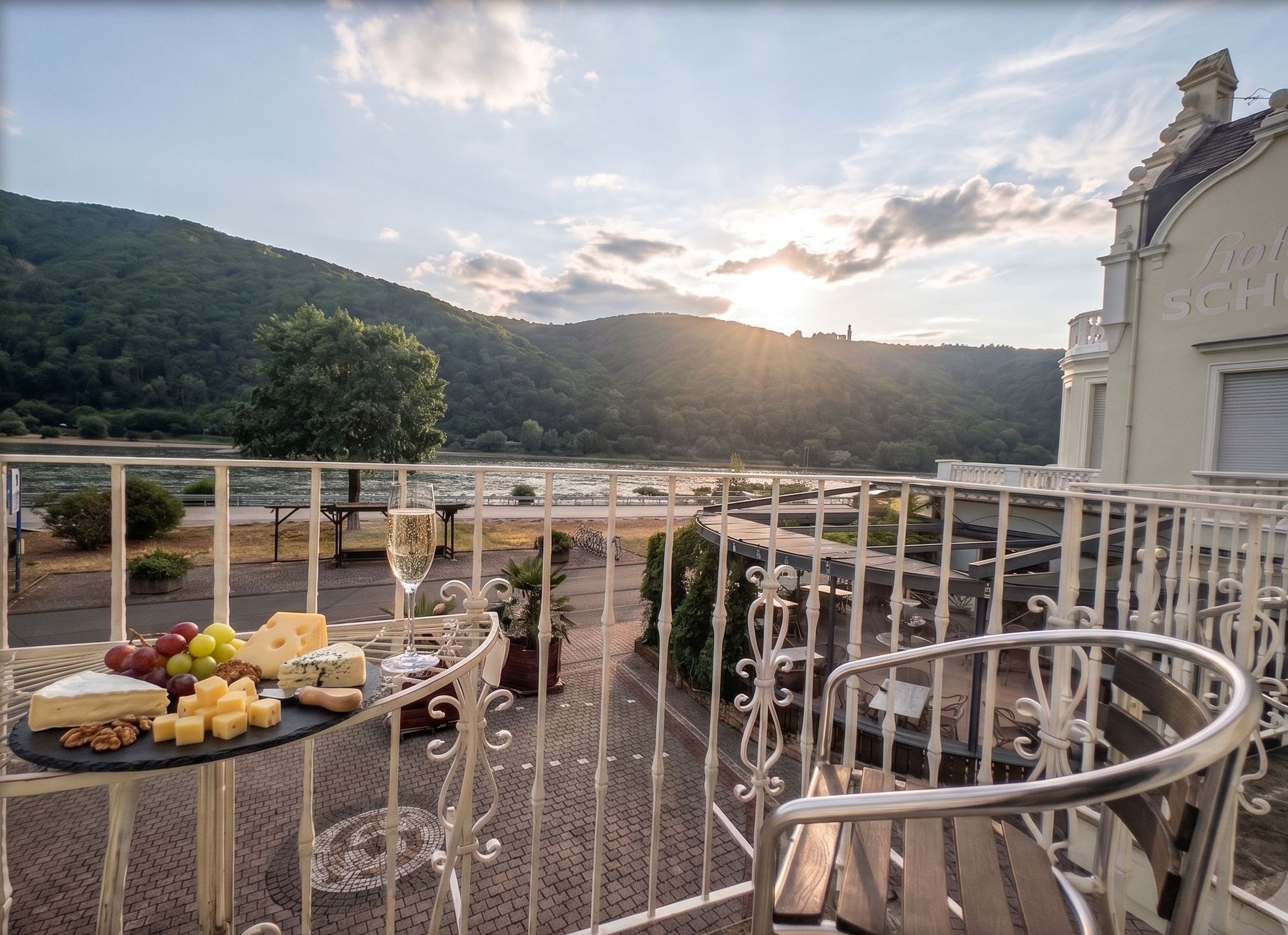 Balkon am Abend mit gedecktem Tischchen, Käse, Trauben und einem Glas Sekt, dahinter der Rhein und die Hügel im Gegenlicht
