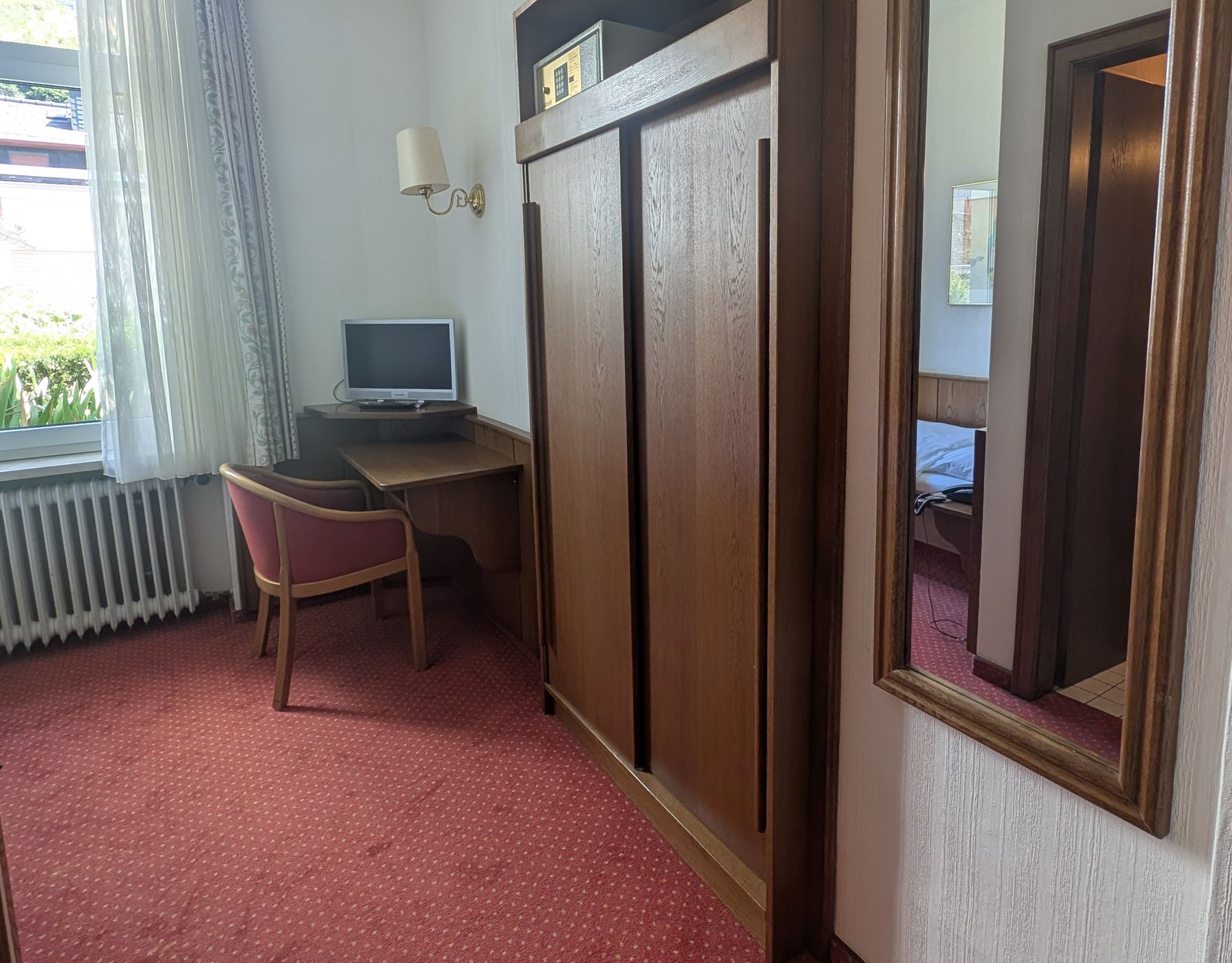 Einzelzimmer mit Schreibtisch, Stuhl, Fernseher und Holzschrank auf rotem Teppichboden