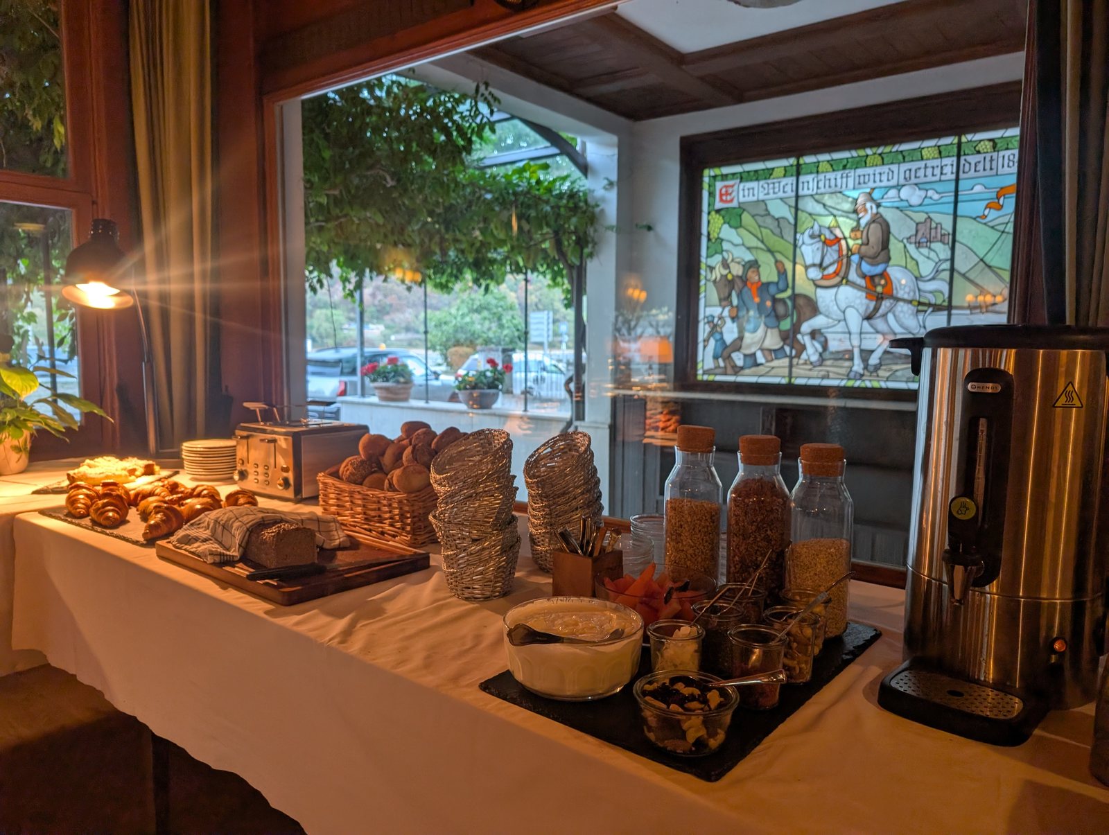 Das Frühstücksbuffet mit Croissants, Brot, Müsli und Joghurt, dahinter das Bleiglasfenster und die Terrasse